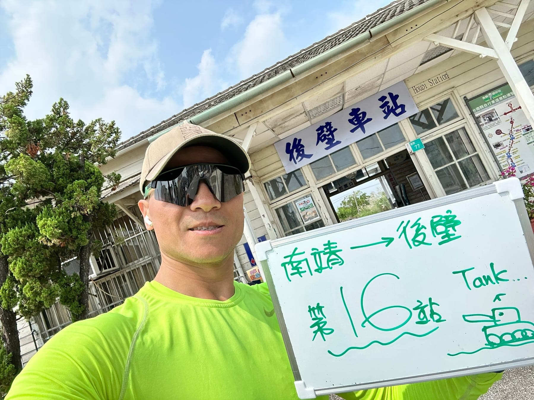 站主在後壁站站牌前的走訪紀錄照，手持寫著「徒步環島 第 16 站　南靖 → 後壁」的白板