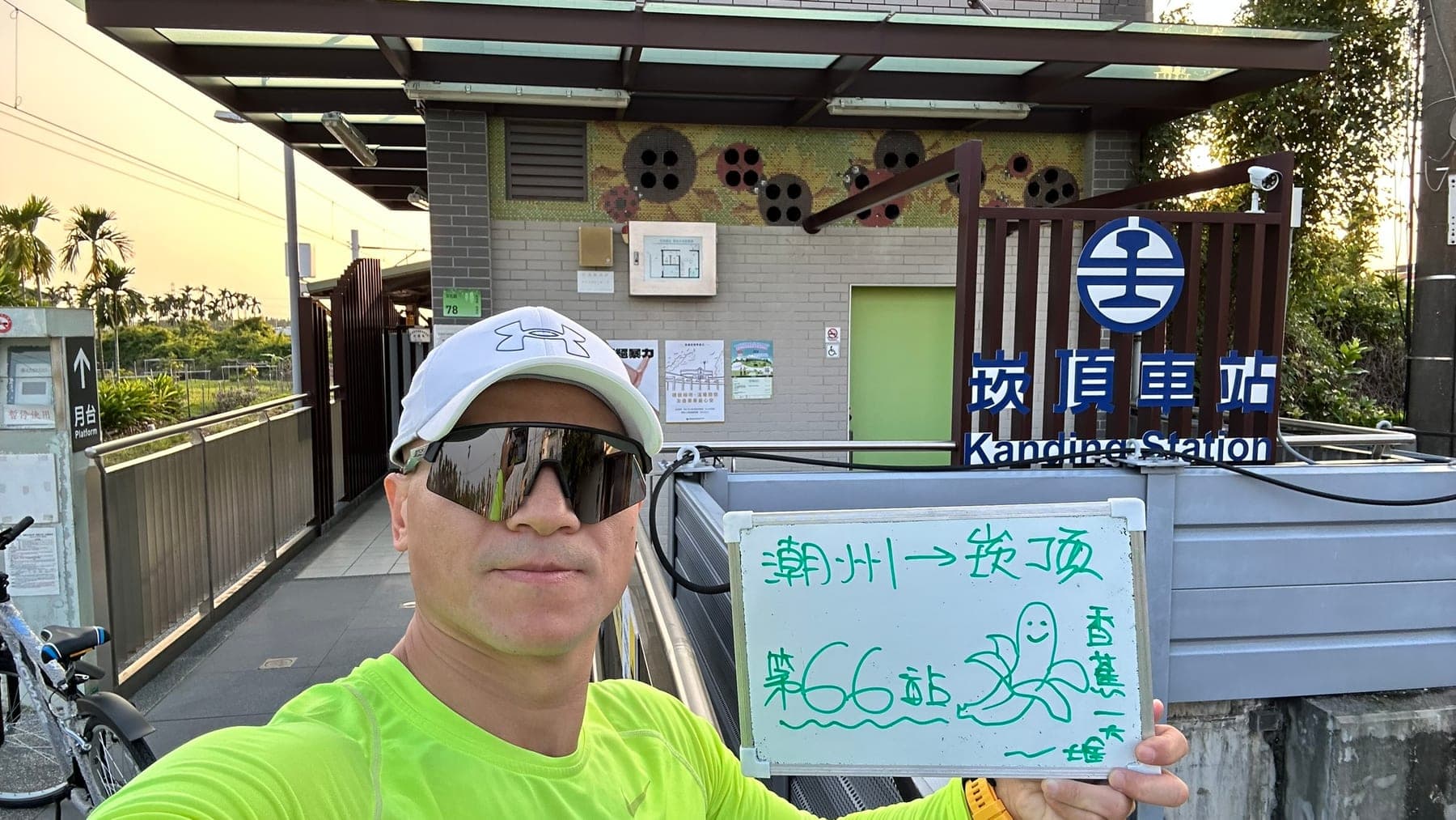 站主在崁頂站站牌前的走訪紀錄照，手持寫著「徒步環島 第 66 站　潮州 → 崁頂」的白板