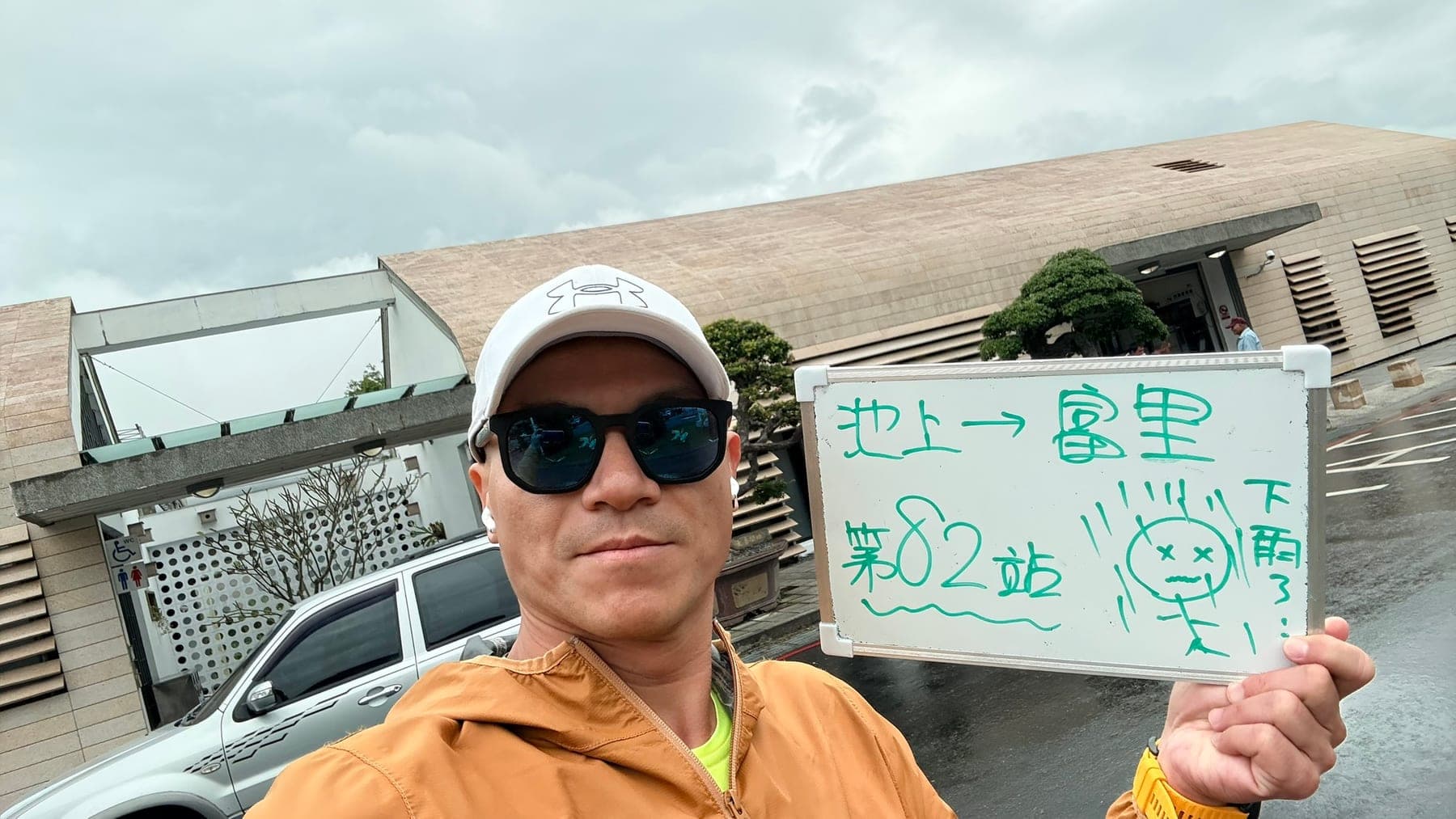 站主在富里站站牌前的走訪紀錄照,手持寫著「徒步環島 第 82 站 池上 → 富里」的白板