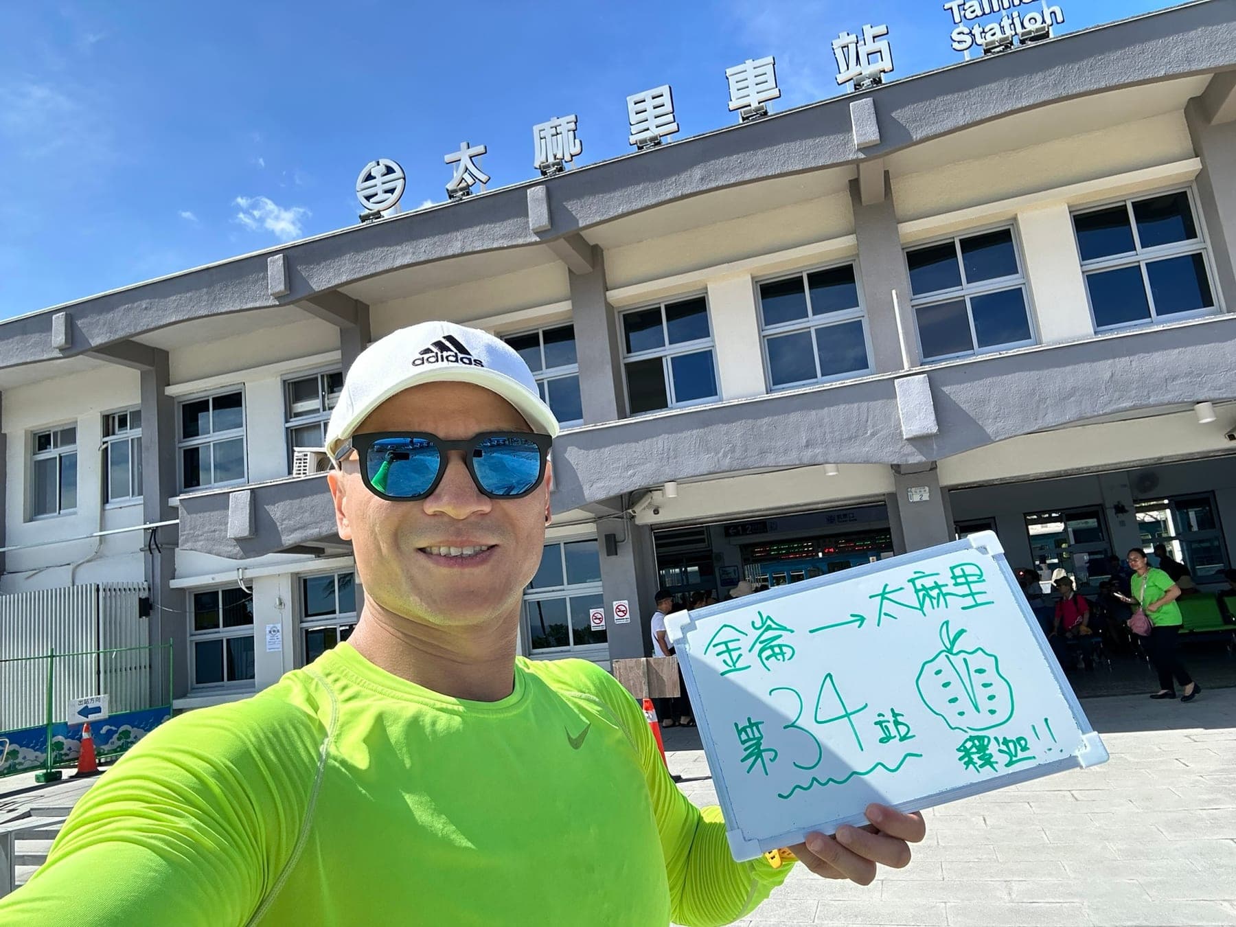 站主在太麻里站站牌前的走訪紀錄照,手持寫著「徒步環島 第 34 站 金崙 → 太麻里」的白板