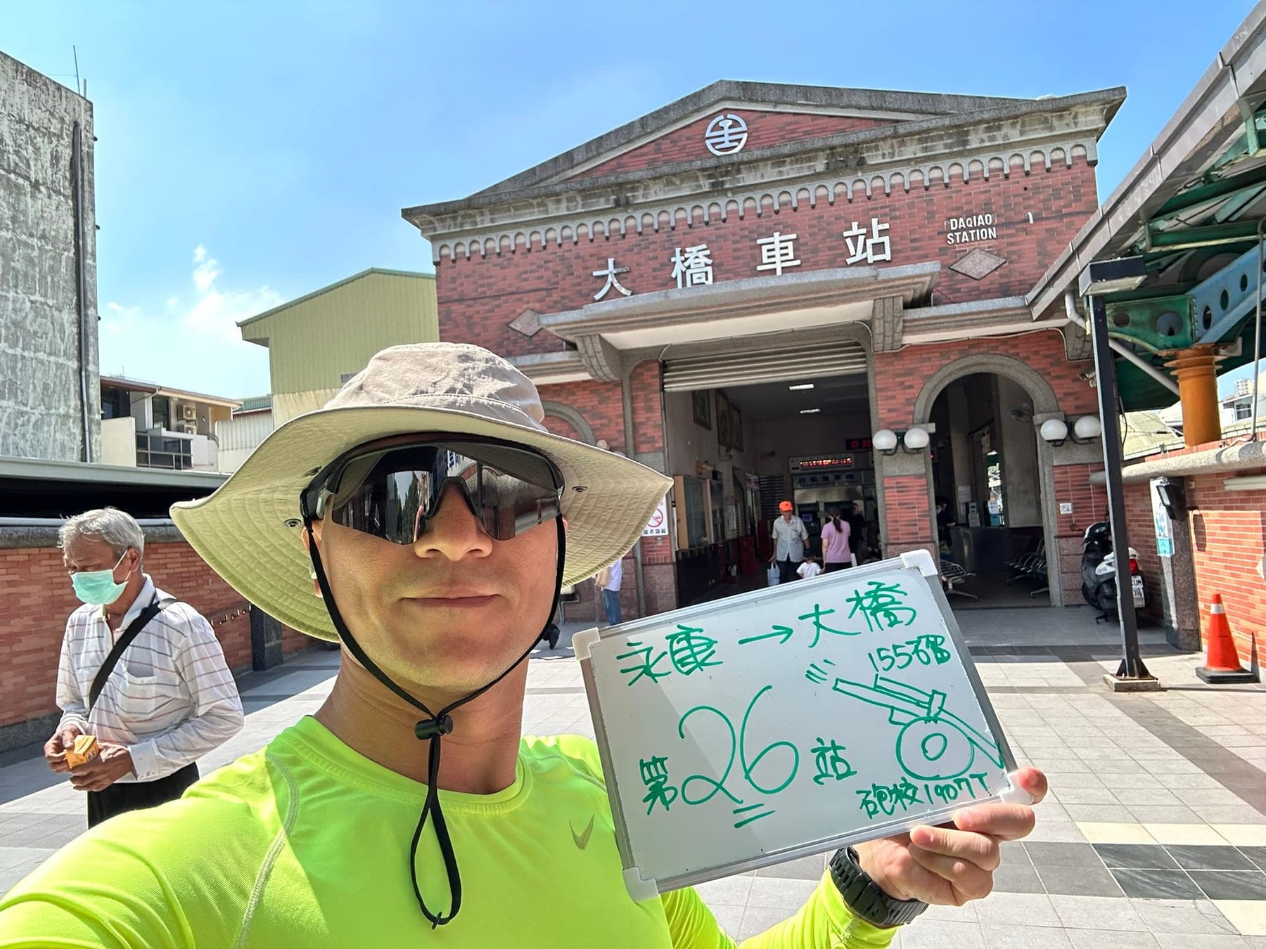 站主在大橋站站牌前的走訪紀錄照,手持寫著「徒步環島 第 26 站 永康 → 大橋」的白板