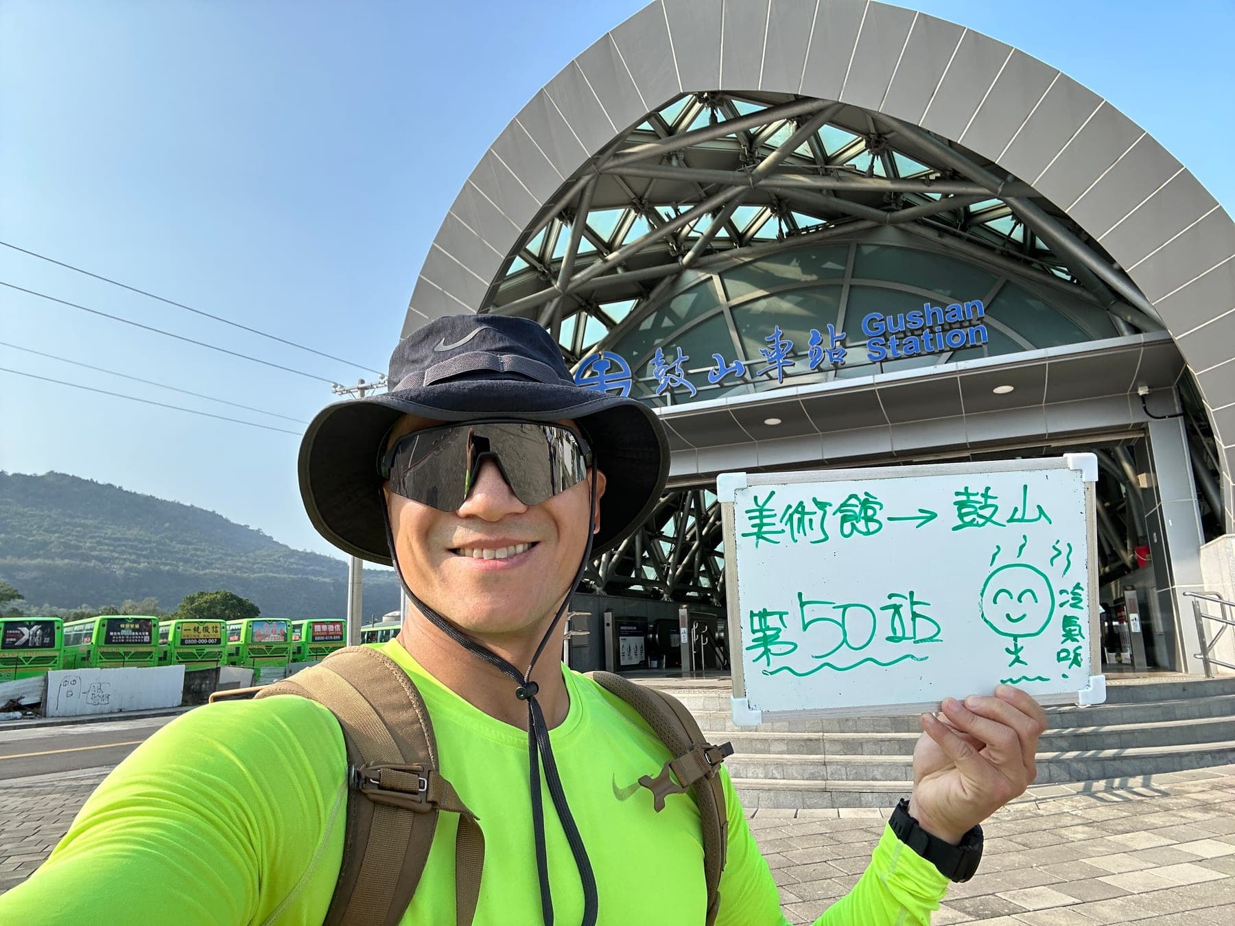 站主在鼓山站站牌前的走訪紀錄照,手持寫著「徒步環島 第 50 站 美術館 → 鼓山」的白板