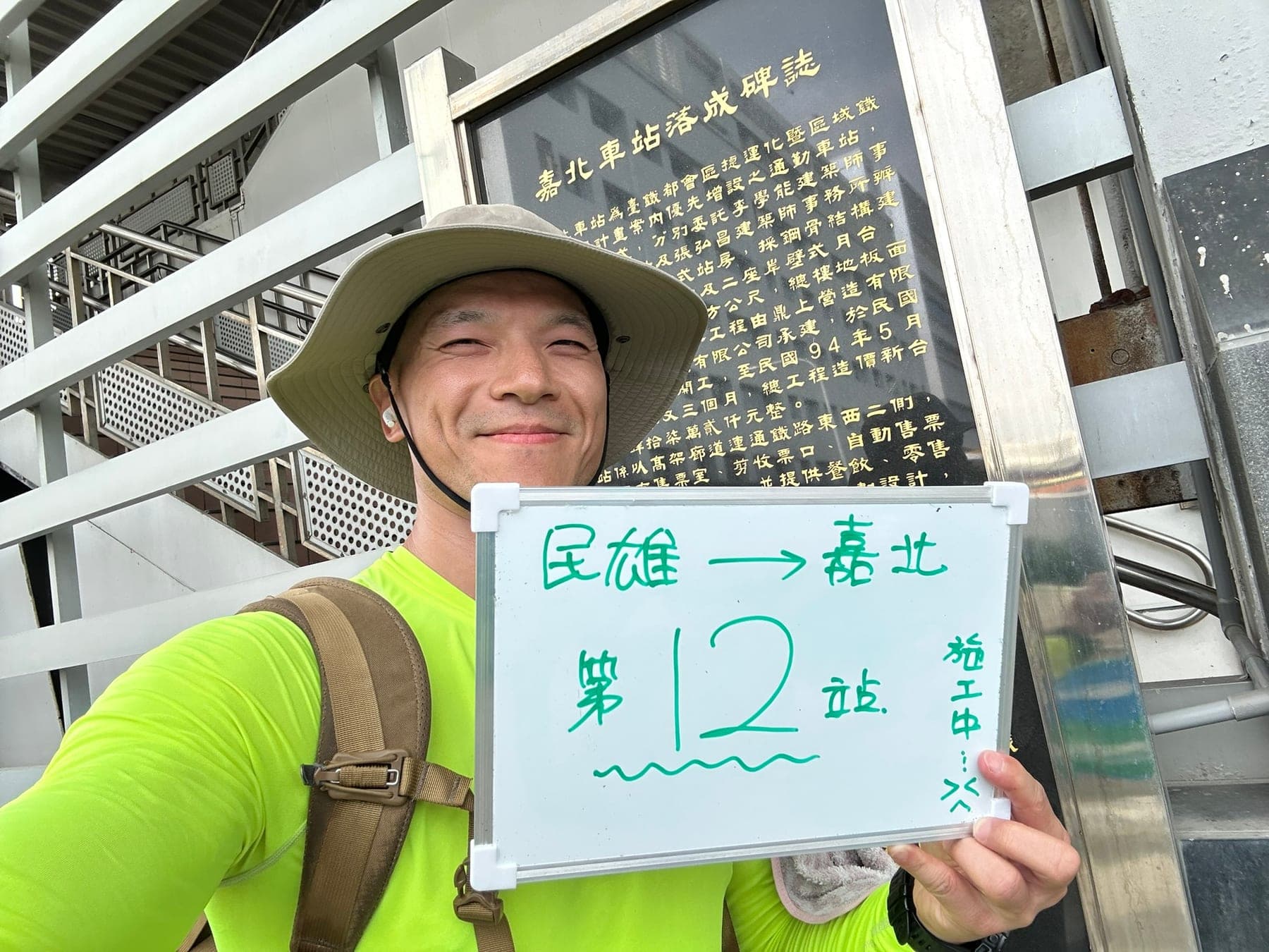 站主在嘉北站站牌前的走訪紀錄照,手持寫著「徒步環島 第 12 站 民雄 → 嘉北」的白板