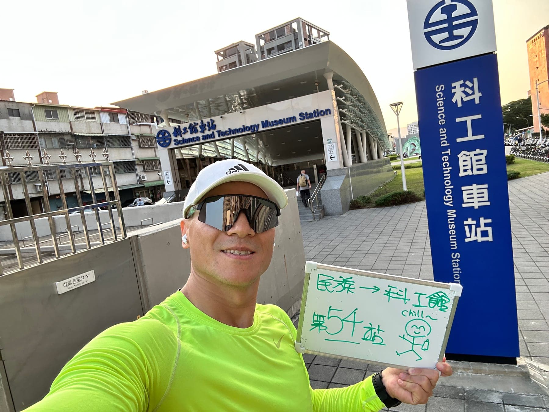 站主在科工館站站牌前的走訪紀錄照,手持寫著「徒步環島 第 54 站 民族 → 科工館」的白板